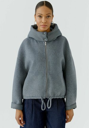 Femme aux cheveux tirés en arrière, portant des boucles d'oreilles anneaux en or, une veste grise zippée avec col rembourré et un pantalon bleu foncé sur un fond uni.