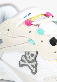 Baskets blanches avec des superpositions en daim beige, des passants de lacets colorés et un emblème brodé de tête de mort et os croisés sur le côté.