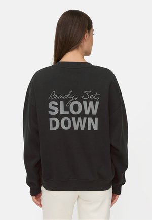 Frau mit langen braunen Haaren, die einen schwarzen Pullover mit dem grauen Schriftzug "Ready, Set, SLOW DOWN" auf dem Rücken trägt und mit dem Rücken zur Kamera steht.