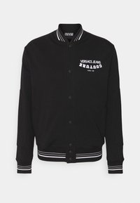 Černá univerzitní bunda z měkké látky, s bílým logem na hrudi, zapínáním na knoflíky a proužkovanými manžetami a lemem.