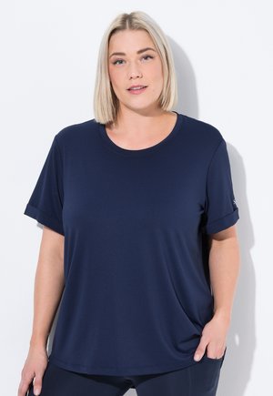Donna bionda con capelli corti che indossa una camicia blu navy a maniche corte e pantaloni coordinati, in posa con una mano in tasca contro un muro bianco.