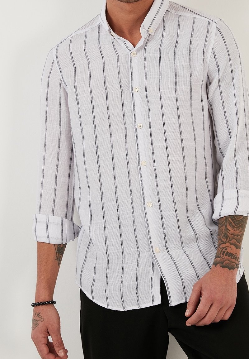 Bílá košile na knoflíky s námořnickými vertikálními pruhy, s ohrnutými rukávy a texturovanou látkou. Má klasický límec a přední zapínání na knoflíky.