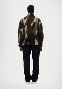 Giacca in pile con un motivo camouflage in verde, marrone e crema. Presenta un colletto alto e una vestibilità comoda. Indossata con pantaloni blu navy.