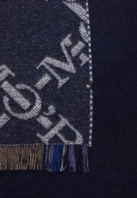 Écharpe en laine bleu foncé avec un grand motif de texte gris clair et une frange multicolore en beige, bleu et violet, sur un fond de tissu foncé.