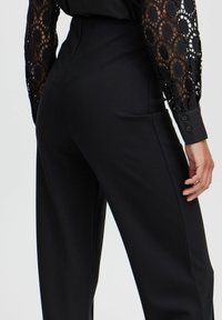 Schwarze Hosen mit taillierter Taille, mit Spitzenverzierungen an den Ärmeln mit kreisförmigen Mustern und knopfbaren Manschetten. Geschmeidige Stofftextur.