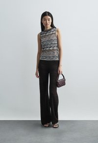 Mönstrad ärmlös topp med vågmönster i flera färger, kombinerad med mörka, texturerade utsvängda byxor; modellen håller en liten vinröd handväska.