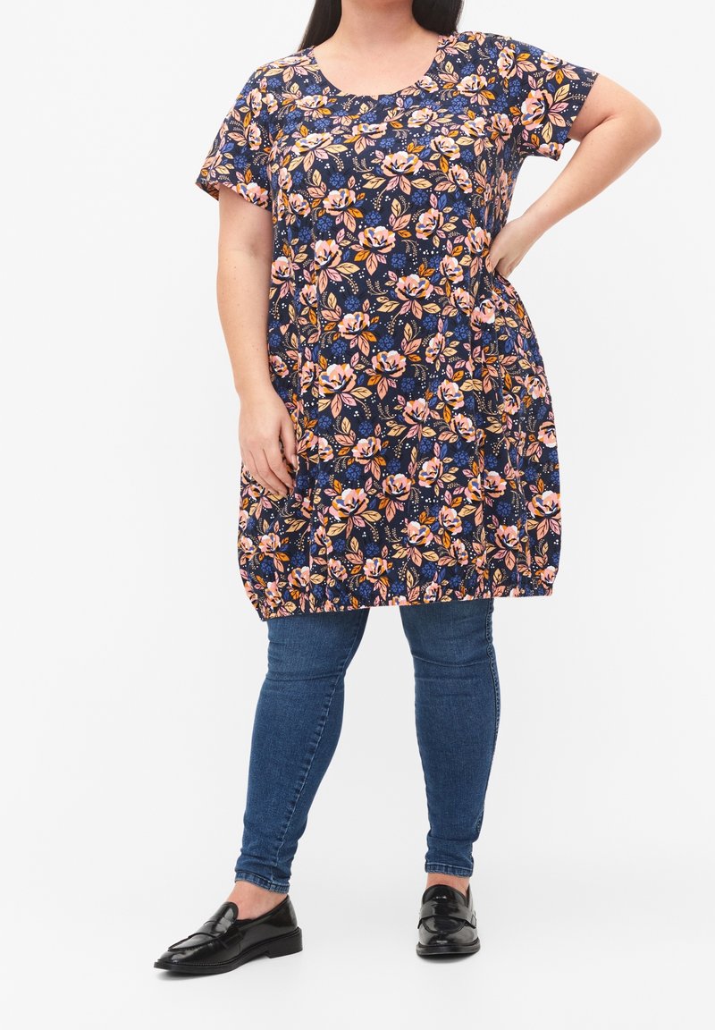 Femme portant une tunique bleu marine à fleurs roses et orange, associée à un jean bleu et des mocassins noirs, debout avec une main sur la hanche.