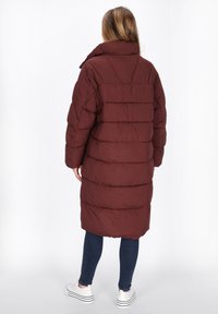 Donna che indossa un lungo cappotto imbottito marrone, jeans blu scuro e sneakers platform bianche, in piedi di spalle su uno sfondo bianco semplice.
