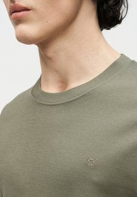 Κοντινή λήψη ενός ατόμου που φοράει ελιάς πράσινη μπλούζα με στρογγυλή λαιμόκοψη και μικρό κεντημένο λογότυπο στο στήθος, εμφανίζοντας το σαγόνι και τον λαιμό.