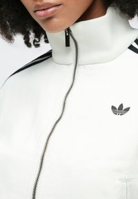 Λευκό μπουφάν με φερμουάρ, ψηλός γιακάς, ριμπ υφή και μαύρες ρίγες στα μανίκια. Διαθέτει μαύρο κεντημένο λογότυπο στο στήθος.