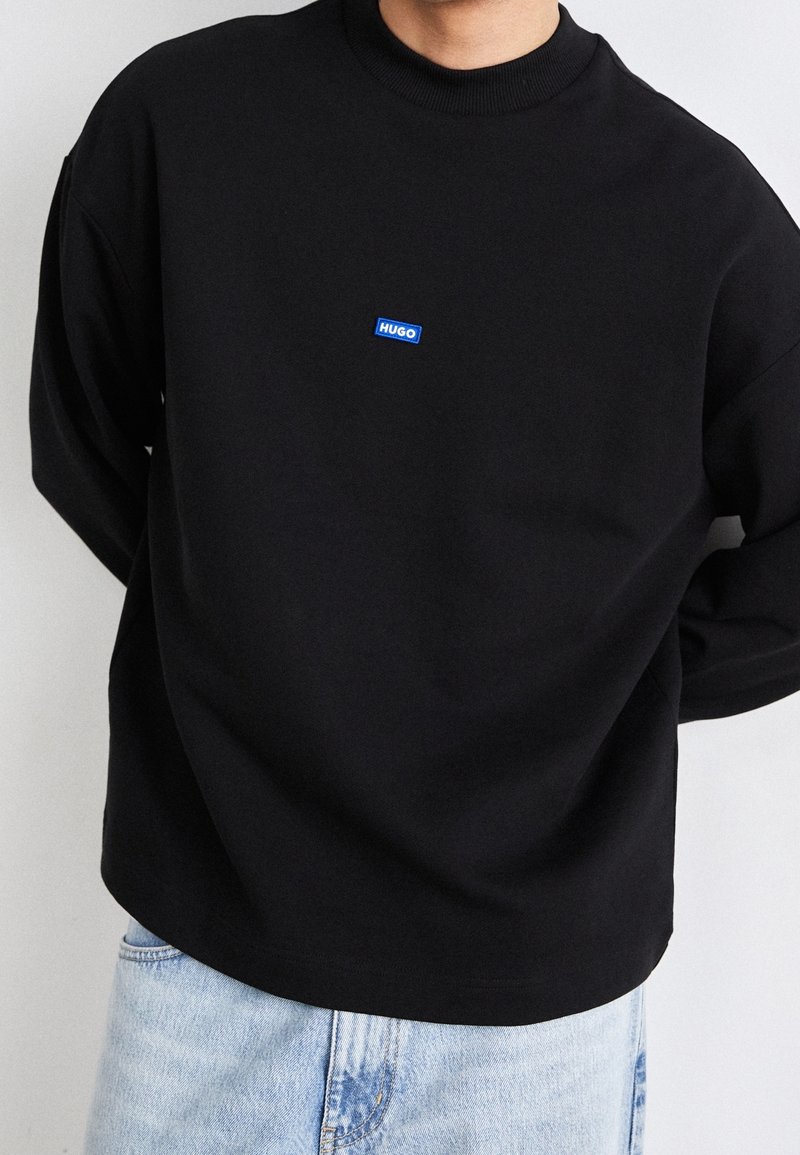 Persona con un sudadera negra de manga larga con un pequeño logo azul "HUGO" en el pecho y jeans de color azul claro.