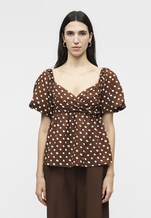 Γυναίκα με μακριά σκούρα μαλλιά που φοράει χρυσούς κρίκους στα αυτιά και καφέ μπλούζα με κοντά μανίκια, λευκές πουά και φαρδύ ντεκολτέ.