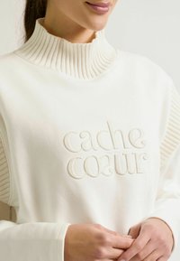 Witte pullover met geribbelde hoge hals en zijdetails, voorzien van reliëf "cache coeur" tekst. Gladde textuur met een lichte glans.