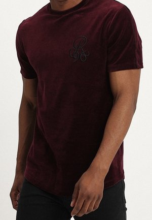 Mężczyzna ubrany w ciemnoczerwoną aksamitną koszulę z krótkim rękawem z czarnym haftowanym logo na lewej piersi, w połączeniu z czarnymi spodniami.