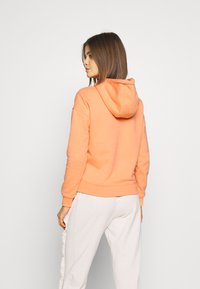 Mikina s kapucňou v svetlo oranžovej farbe, so voľným strihom, rebrovanými manžetami a predným klokankovým vreckom, skombinovaná so svetlými nohavicami.