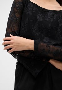 Μαύρη μπλούζα με λουλουδάτο δαντελένιο μοτίβο, διαθέτει μακριά διάφανα μανίκια και στρογγυλή λαιμόκοψη. Το ύφασμα έχει υφασμένη εμφάνιση και επιπλέον λεπτομέρειες.