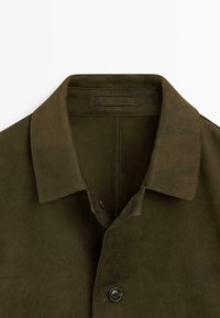 Olivegrüne Wildlederjacke mit spitzem Kragen, einer einzelnen Knopfverschluss und sichtbaren Nähten an den Revers und am Rücken.
