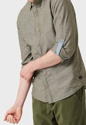 Mann krempelt den Ärmel eines langärmeligen olivgrünen Hemdes mit hellblauem Knopfband hoch, trägt olivgrüne Hose.
