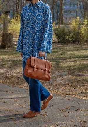 Woman walking outdoors wearing a blue floral jacket and jeans, carrying a brown leather satchel with matching loafers.