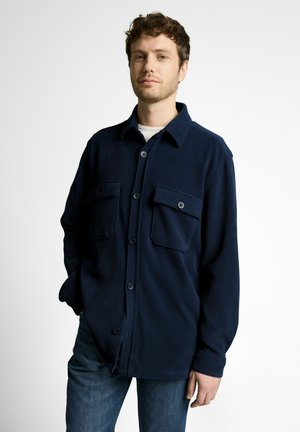 Junger Mann mit hellem Bart, der eine dunkelblaue, mit Knöpfen versehene Hemdjacke mit Klappentaschen und blaue Jeans vor weißem Hintergrund trägt.