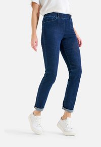 Jeans in denim blu scuro con vestibilità slim e orli risvoltati, dotati di tasche anteriori e dettagli di cucitura chiara, abbinati a sneakers bianche.