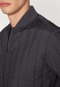 Giacca bomber nera trapuntata con zip frontale, colletto a coste e strisce orizzontali testurizzate. Realizzata in tessuto leggero e lucido.