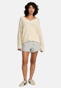 Beiger Strickpullover mit V-Ausschnitt und strukturiertem Rautenmuster, kombiniert mit blau-weiß karierten Shorts. Sneakers vervollständigen das Outfit.