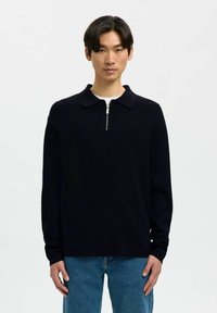 Pullover in maglia blu navy con colletto con zip e maniche lunghe, abbinato a jeans azzurri. Il tessuto sembra liscio e leggermente testurizzato.