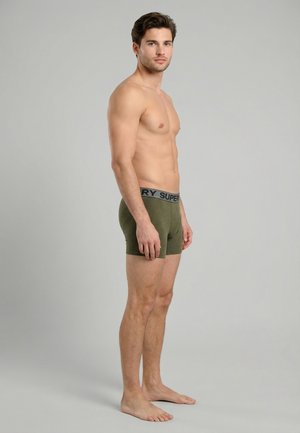 Joven hombre sin camiseta de pie descalzo, con bóxers de color verde oliva con una cinturilla con marca, sobre un fondo gris liso.