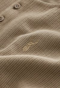 Material textil tricotat de culoare cafenie, cu un model texturat, având două nasturi rotunzi și un design subtil brodat în aur al unei furnici.