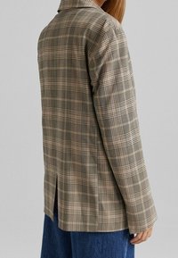 Marynarka w kratę w kolorze brązowym i beżowym, dopasowany krój, z klapami i wentylowanym tyłem. Tkanina ma gładką teksturę i wyrafinowany design.