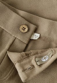 Tessuto di velluto a coste marrone chiaro con chiusura a bottone, cuciture visibili e etichette per abbigliamento. Include un motivo testurizzato e dettagli di accento.