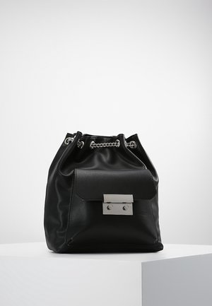 Mochila negra de cuero con cordón y bolsillo frontal asegurado por un broche rectangular plateado, exhibida en un pedestal blanco.