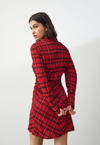 Rotes kariertes Kleid mit langen Ärmeln, hohem Kragen und gerafftem Detail in der Taille. Verfügt über einen kurzen Saum und eine strukturierte Stoffqualität.