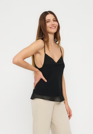Donna con capelli castani lunghi che indossa un top nero senza maniche e pantaloni beige, sorridente, con una mano sul fianco su uno sfondo neutro.