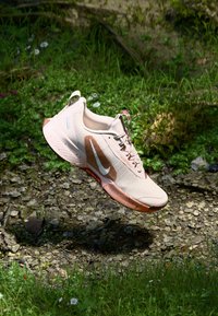 Lys rosa atletisk sko med en tekstureret mesh-overdel, brune accenter, snørebåndsdesign og et hvidt swoosh-logo, svævende over græs og sten.