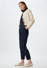 Chaqueta beige corta con botones redondos, pantalones de traje azul marino de tiro alto con pliegues y bailarinas de color crema. Textura suave, diseño a medida.