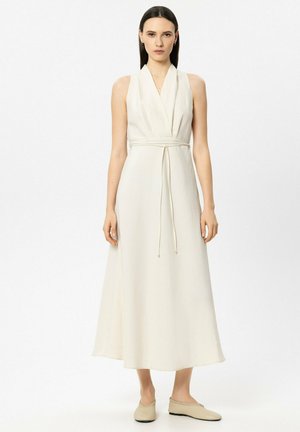 Frau steht in einem ärmellosen, cremefarbenen Midikleid mit V-Ausschnitt und dünnem Gürtel, trägt dazu passende Slipper, vor einem einfarbigen weißen Hintergrund.