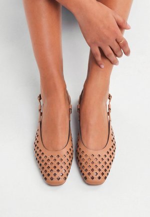 Pies con zapatos planos slingback de cuero trenzado en color beige, una mano apoyada en el tobillo, fotografiados desde arriba sobre un fondo blanco.