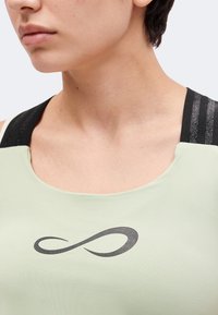 Gros plan d'une personne portant un haut vert clair avec un symbole infini noir sur la poitrine et des bretelles texturées noires.