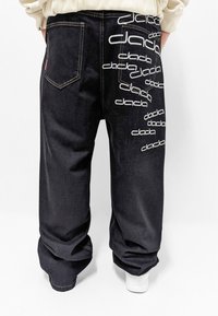 Pantalones de mezclilla negros con un gran estampado blanco del logo "dada" en la parte posterior. Cuenta con dos bolsillos traseros y un corte holgado en los tobillos. Zapatillas blancas visibles.