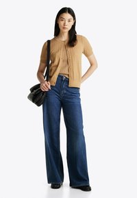 Cardigan de punto beige sobre una blusa beige, combinado con Jeans de pierna ancha de talle alto en azul y un bolso negro de hombro.