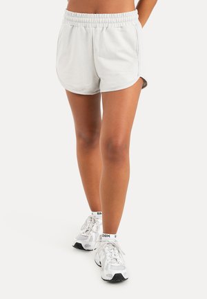 Vrouwelijke benen die witte sportshorts, witte sokken en witte hardloopschoenen dragen, staand op een effen lichte achtergrond.