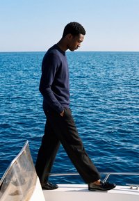 Homme en pull sombre et pantalon marchant sur le bord d'un bateau avec de l'eau océanique en arrière-plan sous un ciel dégagé.