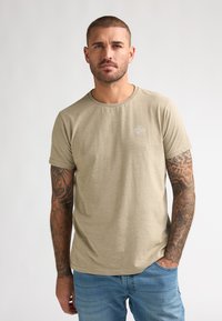 T-shirt beige à manches courtes avec un col rond et des détails de logo subtils, fabriqué en tissu doux, associé à un jean bleu clair.