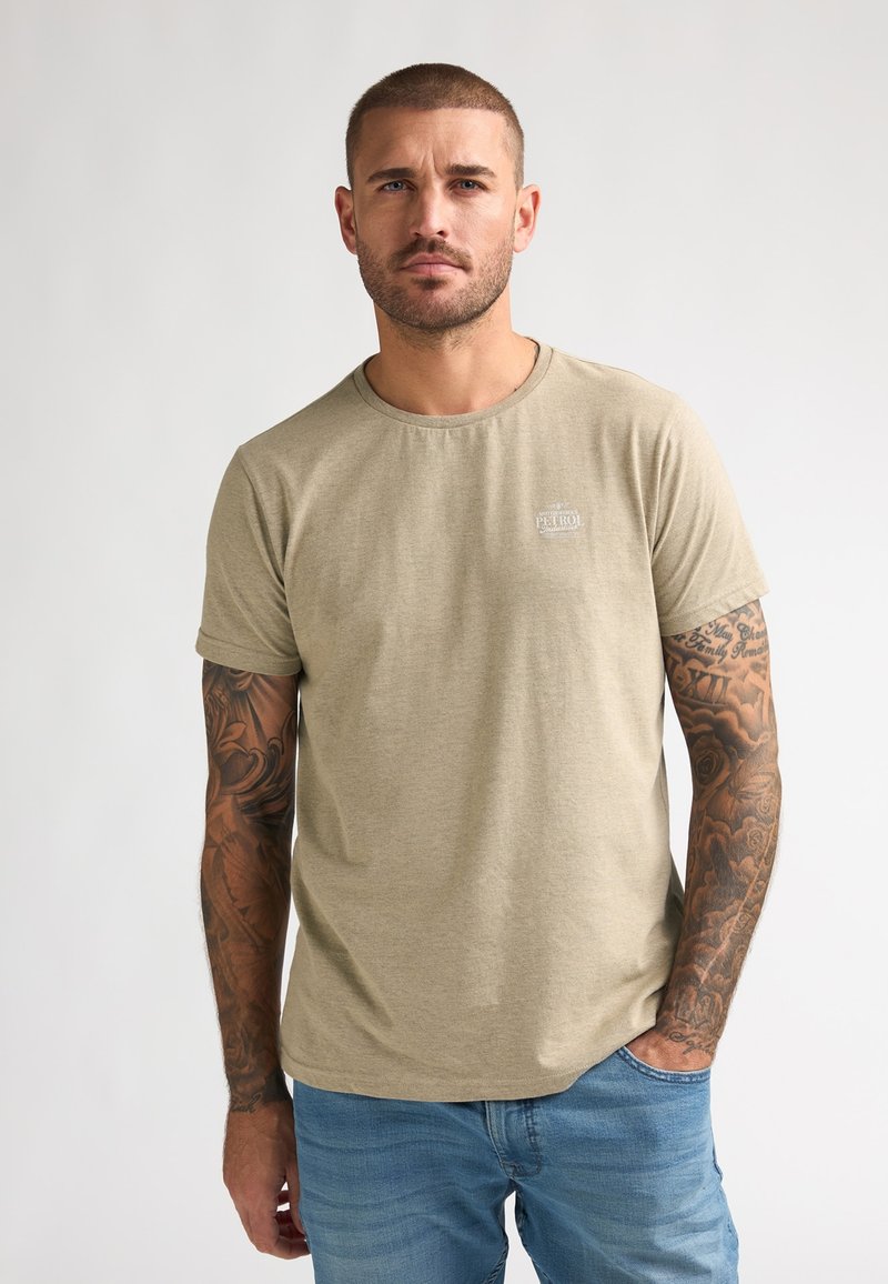 T-shirt beige à manches courtes avec un col rond et des détails de logo subtils, fabriqué en tissu doux, associé à un jean bleu clair.