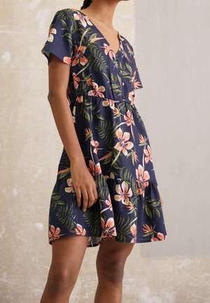Femme portant une robe bleue marine à fleurs avec manches courtes, décolleté en V, devant boutonné et jupe à volants, debout contre un mur texturé.