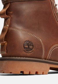 Bota de cuero marrón con marca de impermeabilidad, costuras visibles, suela texturizada y un acento de tela en el costado. Presenta un diseño robusto.