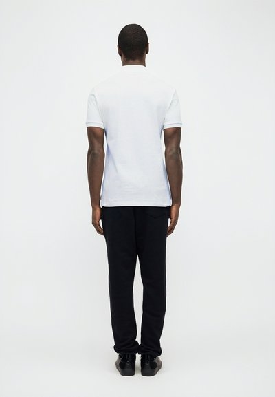 Άνδρας στέκεται με πλάτη προς το φακό, φορώντας λευκό κοντομάνικο πόλο πουκάμισο, μαύρο παντελόνι και μαύρα αθλητικά παπούτσια, μπροστά σε απλό λευκό φόντο.