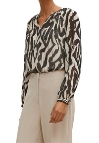 Blouse noire et crème à motifs avec manches longues, décolleté en V, et détails froncés aux épaules, accompagnée d'un pantalon beige.